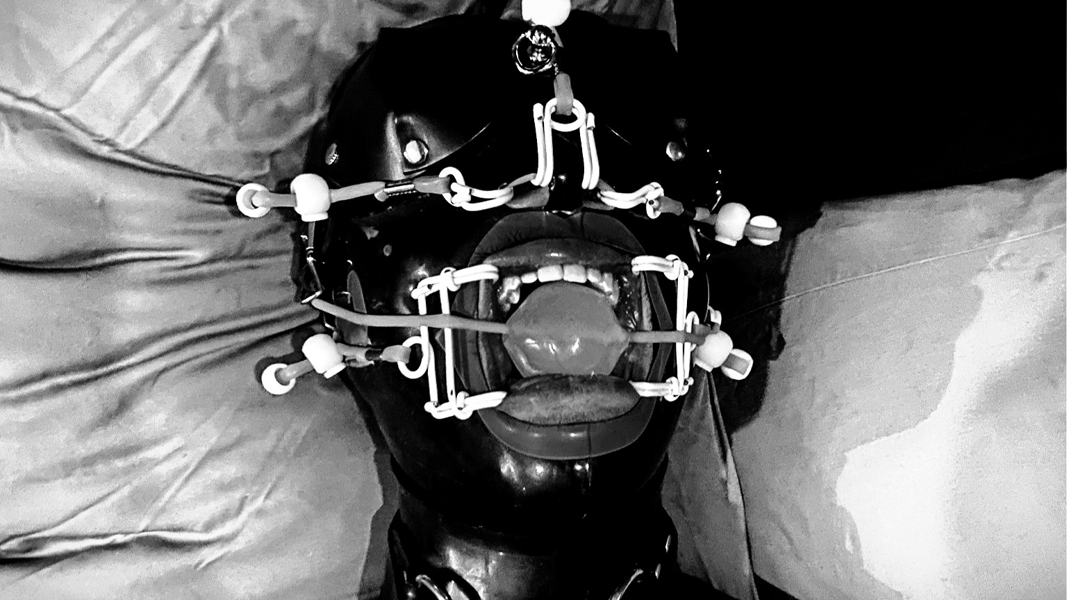 Close-up photograph of Mistrix Violeta's submissive wearing the face hugger gag. The submissive has a black latex hood and a blindfold. The face hugger spreads their nose and mouth apart, and there's a ball gag in the middle of his mouth.