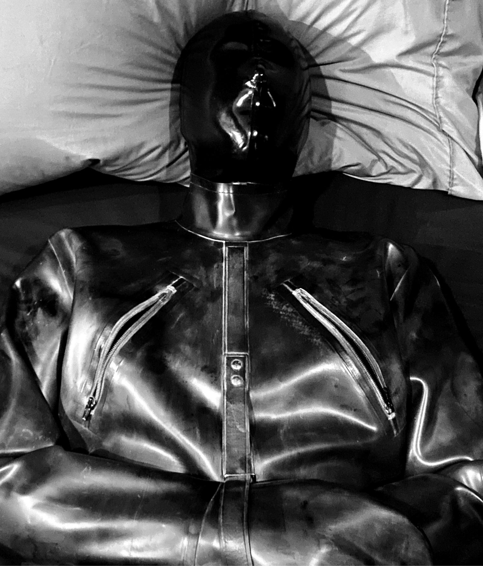Black and white photograph of Mistrix Violeta's submissive in a black latex hood and a latex straight jacket.